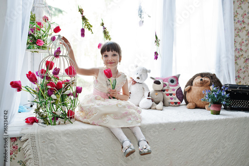 Cute little girl wearing amazing dress sitting on the windowsill next to the flowers with a tupil in her hand.