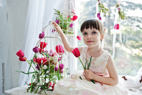 Cute little girl wearing amazing dress sitting on the windowsill next to the flowers with a tupil in her hand. photo