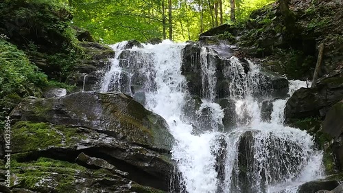 Slow motion shot of a natural waterfall. Waterfall in mountains in slow motion. Close up of a small waterfall in a deep forest. Partial view of an impressive waterfall in the background. 