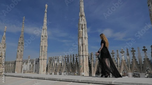Young woman portrait on the rooftop of the famous Duomo cathedral in Milan. Gorgeous fashion woman with blonde hair in elegant black dress posing to the camera.  Video in slow motion.