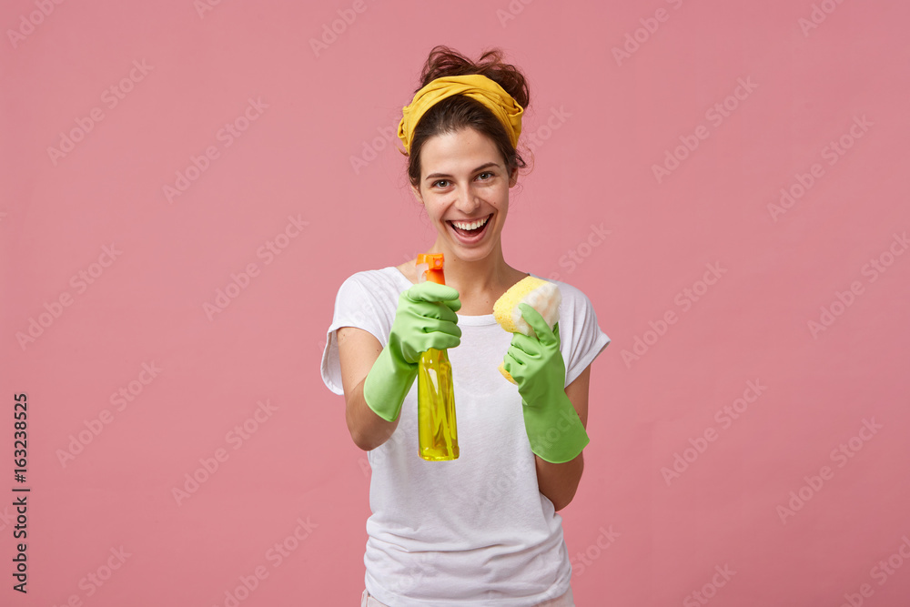 Portrait of happy smiling maid in white tidy T-shirt and green protective gloves demonstrating her detergent and sponge before work. People, housework, housekeeping and housecleaning concept