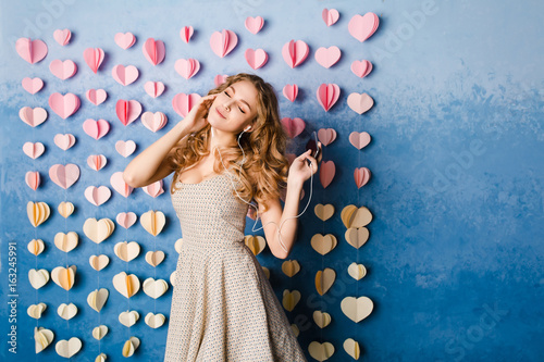 Cute sexy slim girl with blond curly hair standing in a studio with blue background and listenign to music on earphones. She smiles and looks sexy. photo