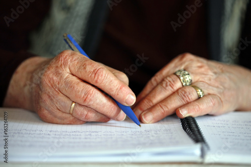 Close up of elderly woman's hands with pen; old woman is writing; life-long learning concept; selective focus background.