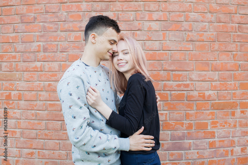 Happy young couple hugging and smiling on a background of red brick wall. Blonde girl with blue eyes and a young man of Arab appearance with brown eyes