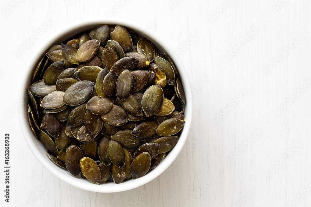 Hulled pumpkin seeds in white ceramic bowl isolated on painted white wood from above. Hulled pumpkin seeds in white ceramic bowl isolated on painted white wood from above.