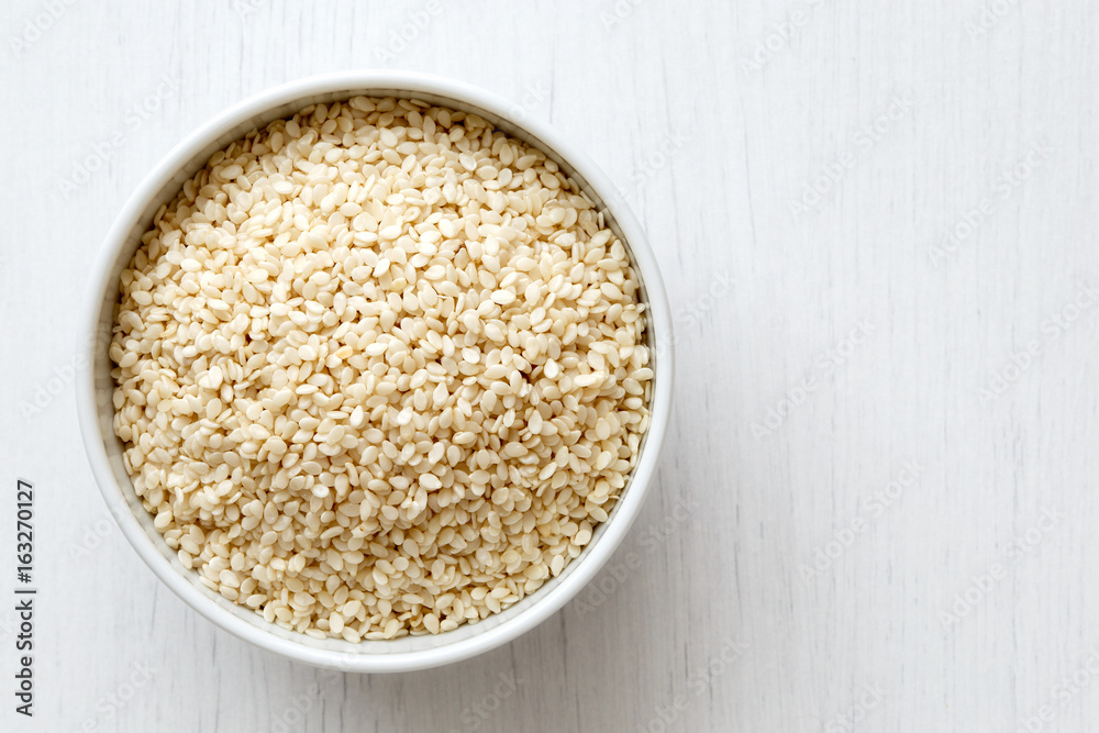 Decorticated sesame seeds in white ceramic bowl isolated on painted white wood from above. Decorticated sesame seeds in white ceramic bowl isolated on painted white wood from above.