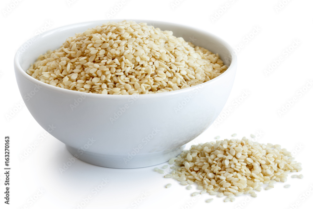 Decorticated sesame seeds in white ceramic bowl isolated on white. Spilled seeds. Decorticated sesame seeds in white ceramic bowl isolated on white. Spilled seeds.