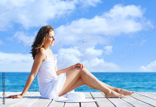 Beautiful, attractive model posing in white dress on a wooden pier. Sea and sky background. Vacation, traveling and resort concept. © Acronym