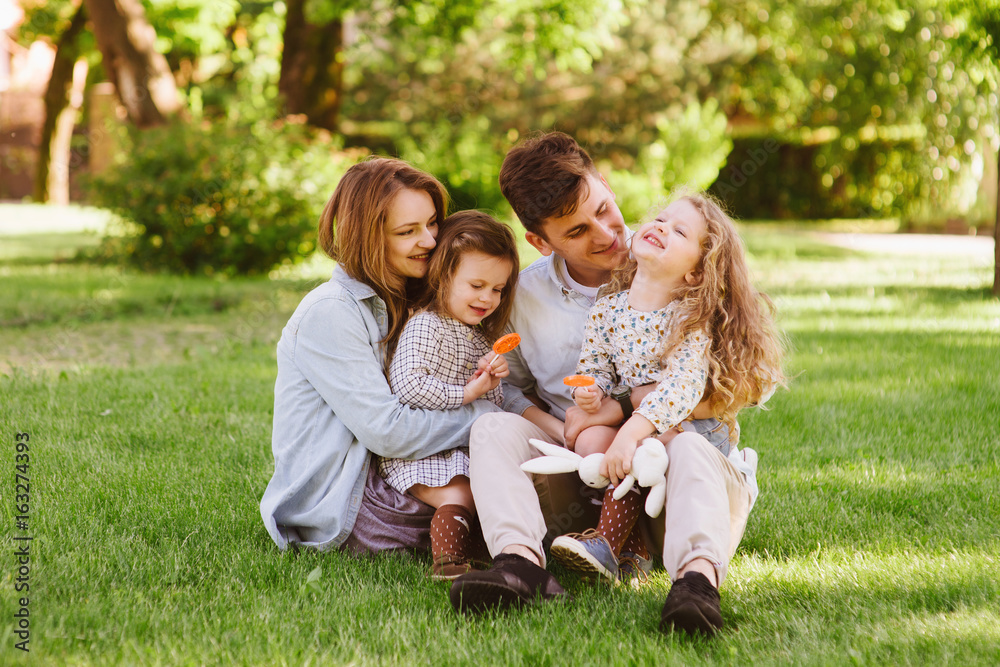 Young cheerful family sitting on grass enjoying vocation and keeping their children on knees. People sitting in summer park. Sunny day, outdoors Young cheerful family sitting on grass enjoying vocation and keeping their children on knees. People sitting in summer park. Sunny day, outdoors