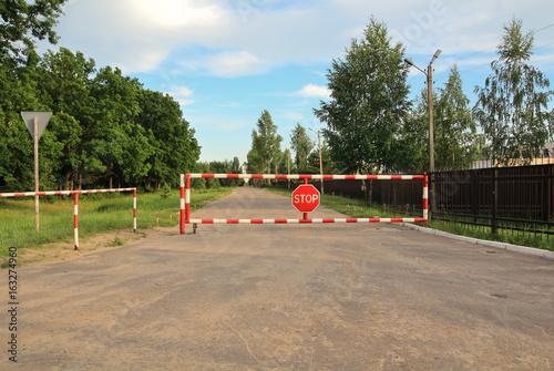 Red and white colored street barrier with sign "stop" on it. Closed Road. Fence around restricted military base area.