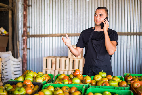 Young man speak on phone with costumers in front collect tomatoes boxes at greenhouse. Online phone sales of tomato  orders of costumers family farm business. photo