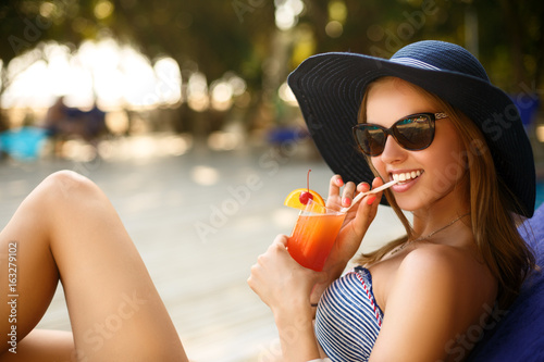 Portrait of young woman with cocktail glass chilling in the tropical sun near swimming pool on a deck chair with palm trees behind. Vacation concept