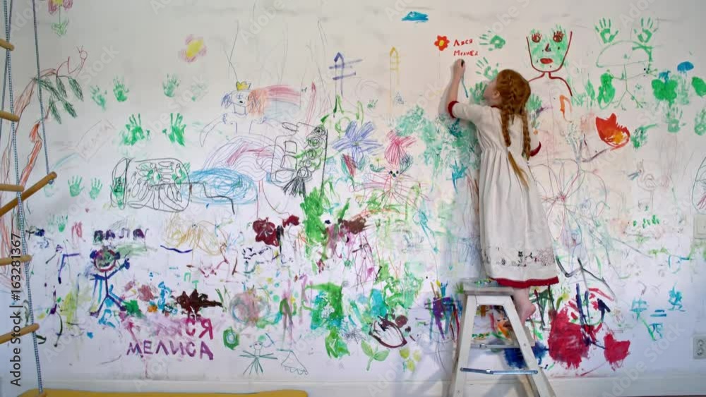 Wide shot of little redhead girl with braids standing on small stepladder and doodling with colored crayon on wall covered in colorful drawings and words