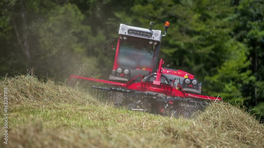 A young man is driving his tractor on a hill and with the help of agricultural machinery he is arranging hay into piles.