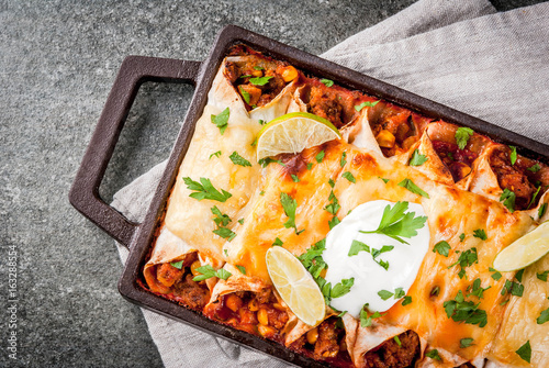 Mexican food. Cuisine of South America. Traditional dish of spicy beef enchiladas with corn, beans, tomato. On a baking tray, on a black stone background. Top view copy space
