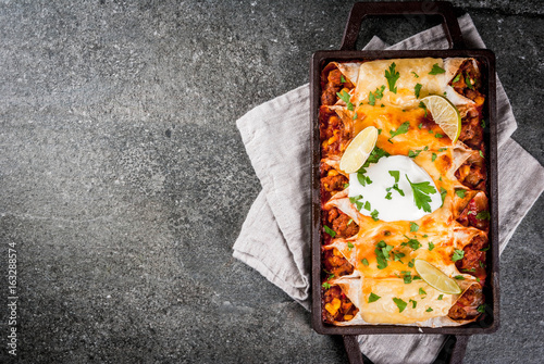 Mexican food. Cuisine of South America. Traditional dish of spicy beef enchiladas with corn, beans, tomato. On a baking tray, on a black stone background. Top view copy space photo