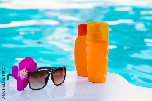 Bottles of tan cream and lotion, sunglasses near swimming pool