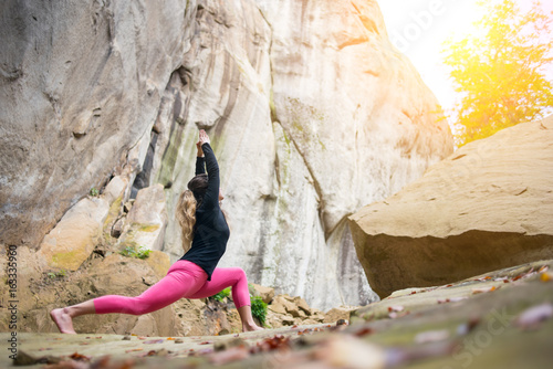 Flexible fit female is practicing yoga and doing asana Virabhadrasana 1 outdoors. Huge rocks on the background