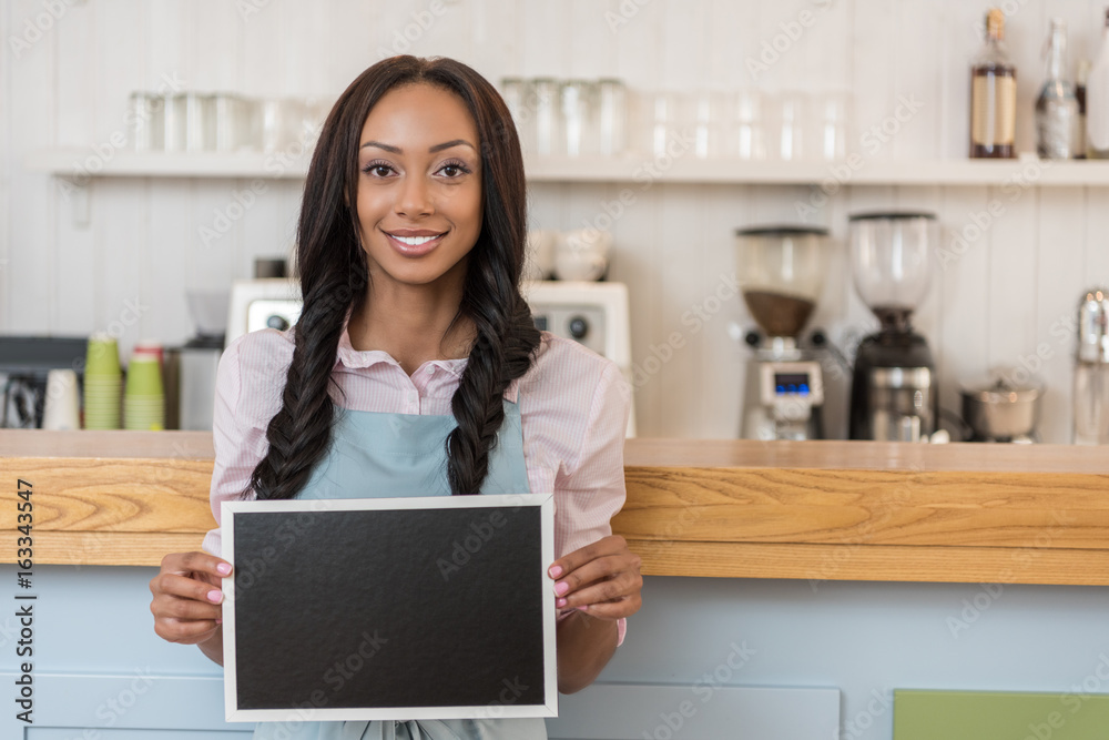 afro-american waitress holding empty board and looking at camera while standing at cafe afro-american waitress holding empty board and looking at camera while standing at cafe
