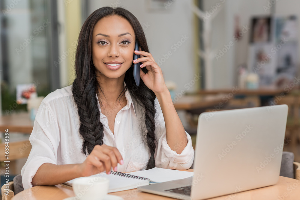 young beautiful african american businesswoman talking on smartphone while sitting near laptop at cafe young beautiful african american businesswoman talking on smartphone while sitting near laptop at cafe