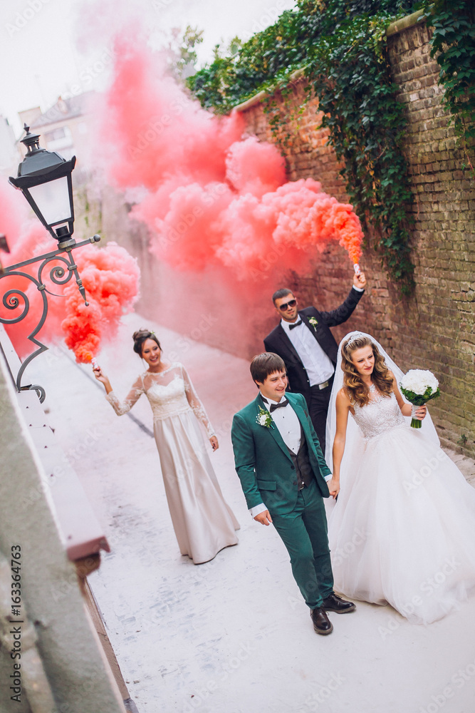 Newlyweds and their friends raise their hands with colors smoke. Running with smoke bombs. Red and blue colors. Wedding couple with color smoke.