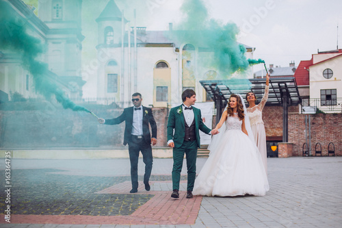 Newlyweds and their friends raise their hands with colors smoke. Running with smoke bombs. Red and blue colors. Wedding couple with color smoke.