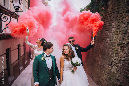 Obraz na płótnie Newlyweds and their friends raise their hands with colors smoke. Running with smoke bombs. Red and blue colors. Wedding couple with color smoke. w salonie