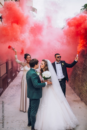 Newlyweds and their friends raise their hands with colors smoke. Running with smoke bombs. Red and blue colors. Wedding couple with color smoke.