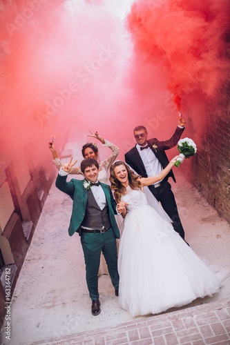 Newlyweds and their friends raise their hands with colors smoke. Running with smoke bombs. Red and blue colors. Wedding couple with color smoke.