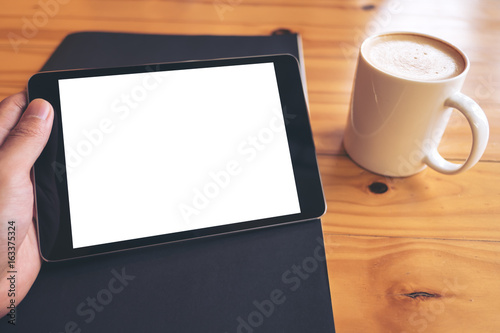 Mockup image of hand holding black tablet pc with blank white screen on a black paper and white coffee cup on vintage wooden table background