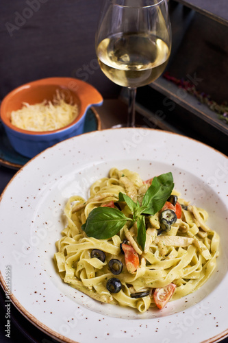 Pasta with chicken, olives and cheese on ceramic plate with glass of white wine on wooden table in rustic style.