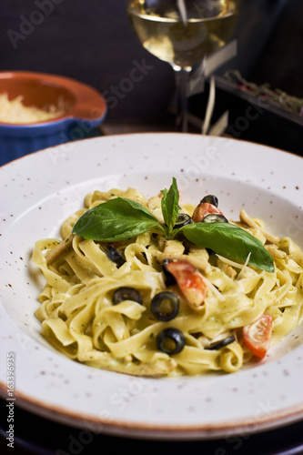 Closeup view of Pasta with chicken, olives and cheese on ceramic white plate with glass of white wine on table