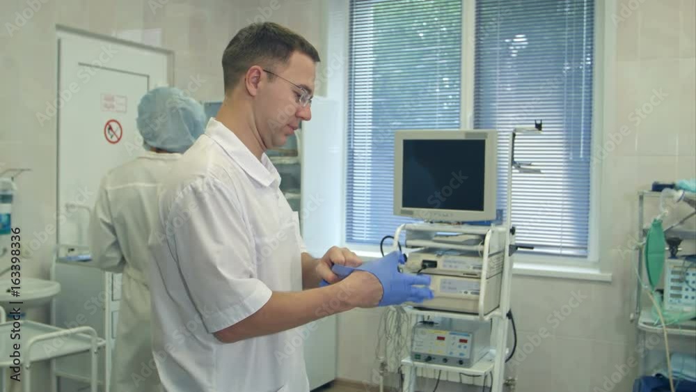 Male surgeon putting on sterile rubber gloves for surgery while nurse washing her hands in a surgery room Male surgeon putting on sterile rubber gloves for surgery while nurse washing her hands in a surgery room