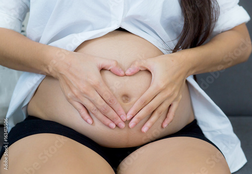 Pregnant asian woman holding gesture heart symbol shape belly with on sofa in home. Maternity concept.