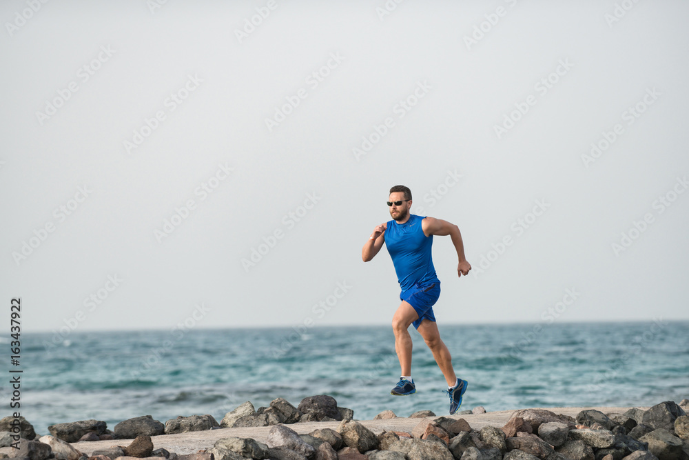 Strong sportsman in blue shorts and shirt jogging along coast