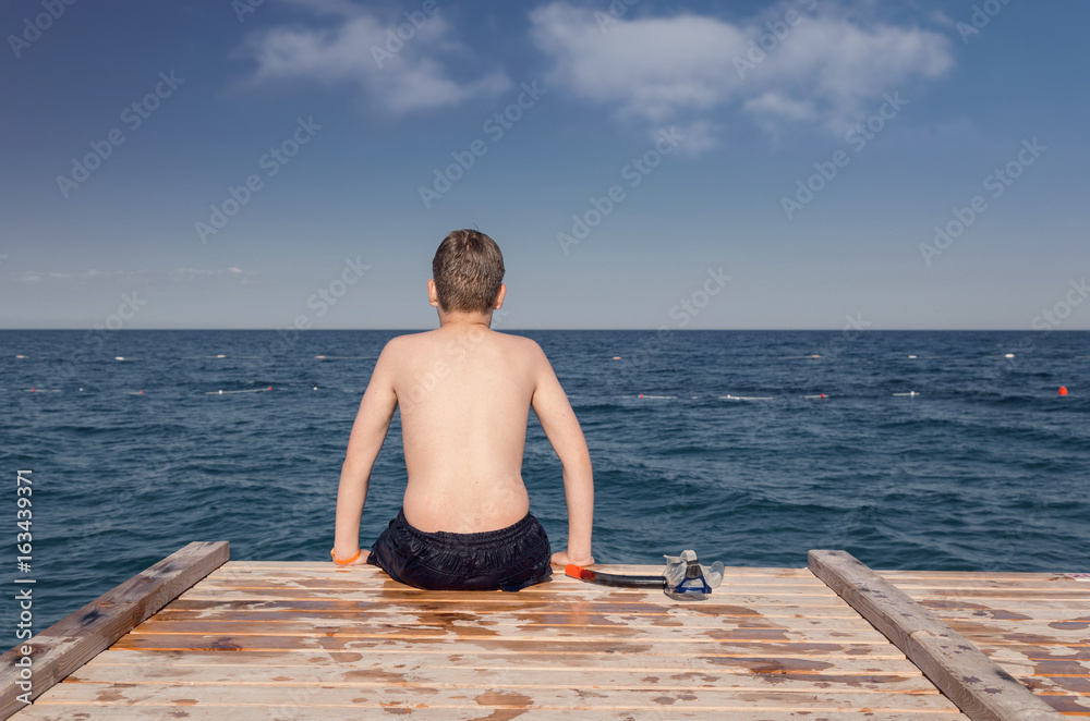 Boy with diving mask, sitting on a wooden jetty at sea beach. Daydreaming concept.