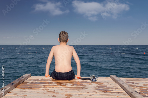 Boy with diving mask, sitting on a wooden jetty at sea beach. Daydreaming concept.