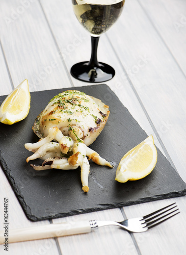 Cuttlefish with garlic and parsley, lemon next to white wine glass on whiteboard. Vertical studio shot.