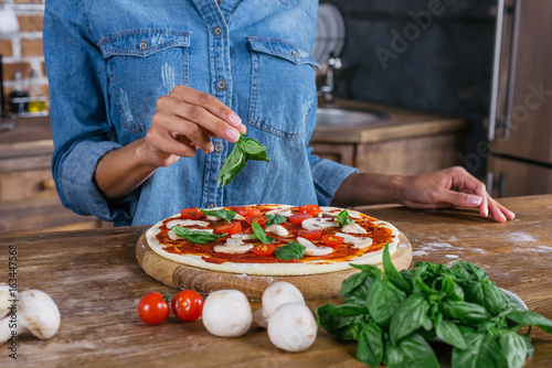 Fototapeta Naklejka Na Ścianę i Meble -  Cropped shot of woman putting basil leaves on raw pizza