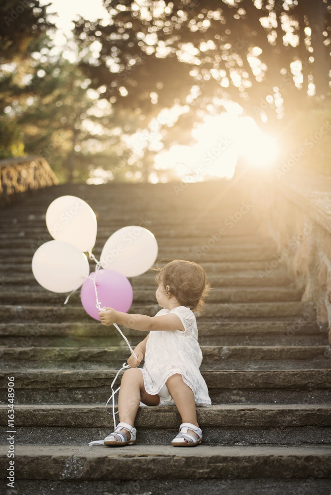 Baby girl standing on stairs and holding balloons. Vintage looking. Sun, sun haze, glare Baby girl standing on stairs and holding balloons. Vintage looking. Sun, sun haze, glare