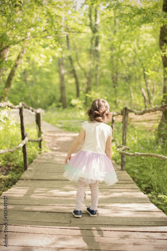 A little girl in back with a tutu dress on a wooden bridge in a forest. Wooden walkway in green forest near the Ropotamo river, Bulgaria