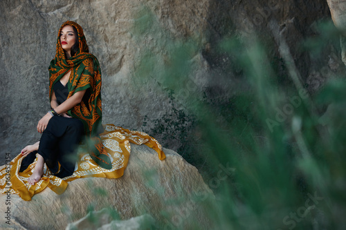 Beautiful Muslim woman with oriental scarf on head sitting on cliff on a mountain background
