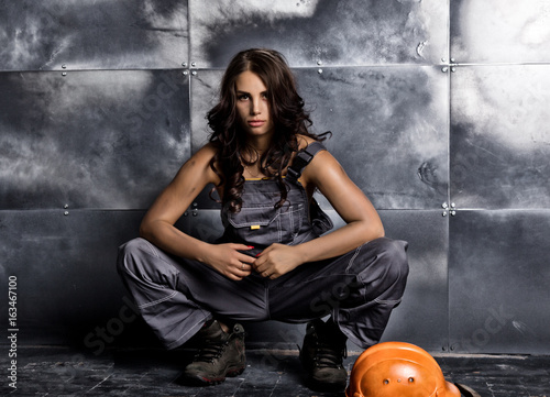 Beautiful sexy worker with a pickaxe and helmet. Seductive and beautiful woman miner sitting on a floor on steel background