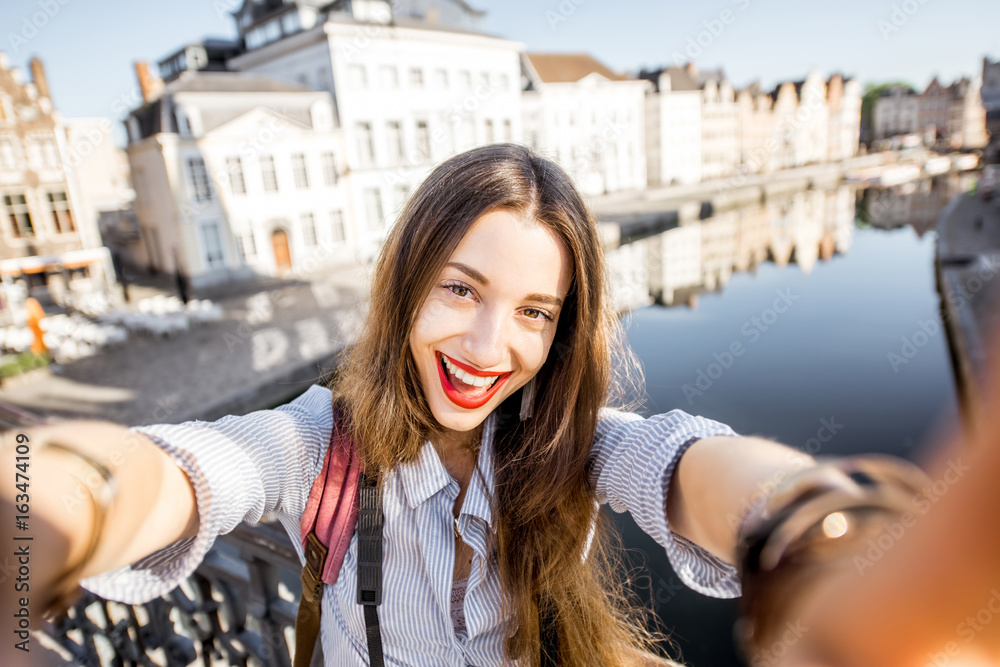 Young woman tourist making selfie photo standing on the bridge with beautiful view on Gent city in Belgium