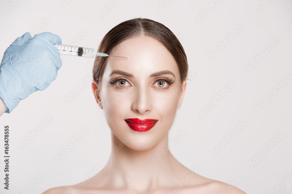 Portrait of beautiful happy young woman ready anti aging serum shot in forehead wrinkle area plastic surgery doctor's hand in blue gloves holding injection over woman's face. Positive human expression