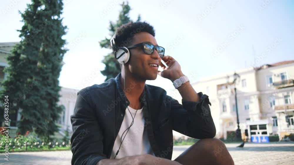 Portrait of young stylish hipster black man in white headphones and sun glasses dancing outdoor in city centre, having some fun Portrait of young stylish hipster black man in white headphones and sun glasses dancing outdoor in city centre, having some fun
