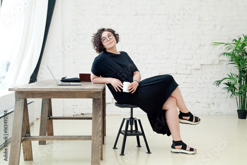 Young plus size female model sitting in front of the table and working on her notebook. Bright modern coworking hall. Composition with copy space. photo