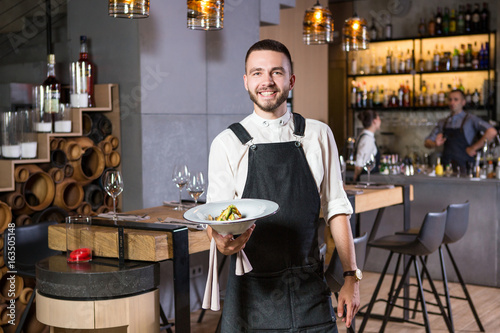 A handsome young guy with a beard dressed in an apron standing in a restaurant and holding a white plate with a moth. Against the background, the bar counter and loft style interior photo