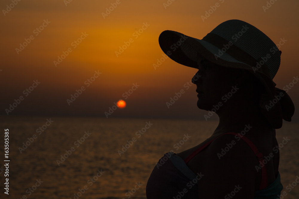 Woman silhouette with hat sitting on sunset sea background, back lit,Silhouette of woman in hat on colorful and vivid sunset background
