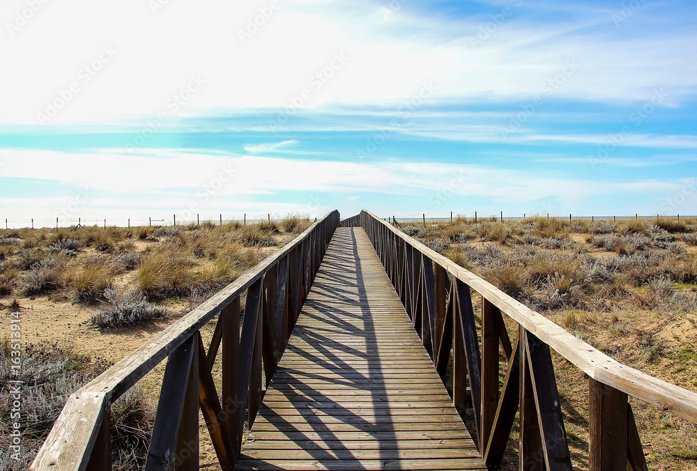 Wooden walkway over the sand dunes to the beach. Beach pathway in Huelva Beach, inside a natural area in Andalusia, Spain with chemical industries in background